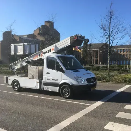 Een wit busje met een kraan erop rijdt door een straat.