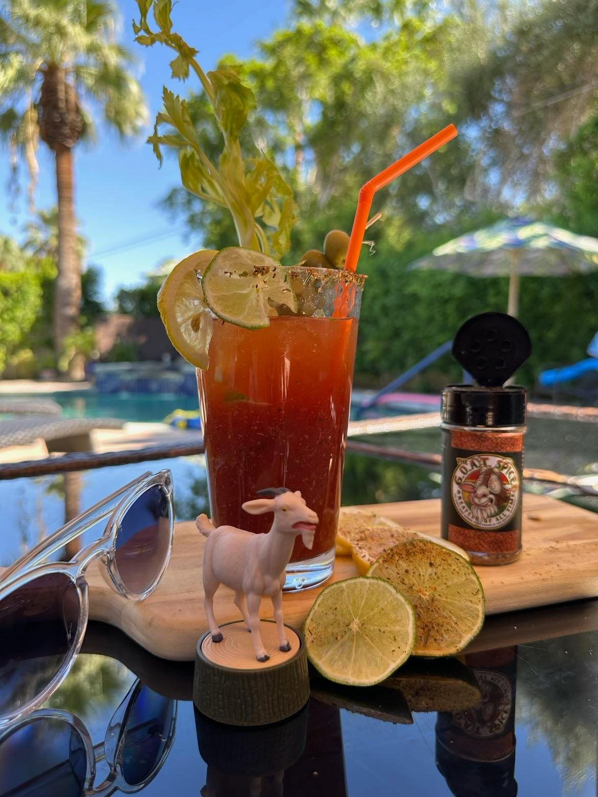 Tall red cocktail with lime and straw on a poolside table beside sunglasses and a spice shaker.