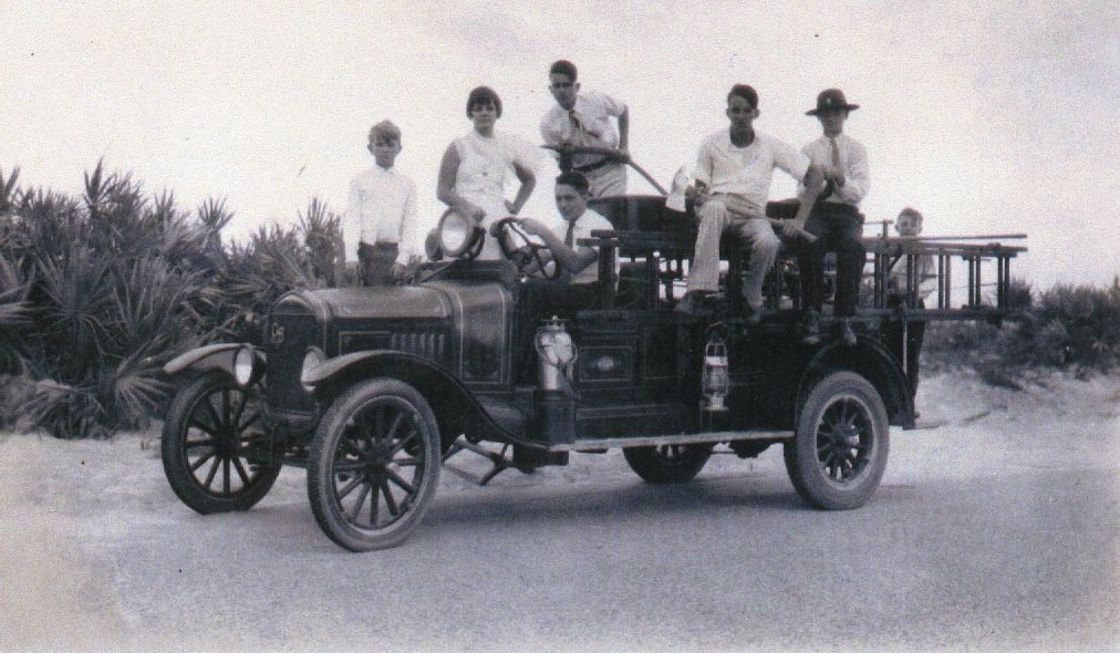 Vintage open-top truck with six people standing on it, parked on a dirt road near bushes