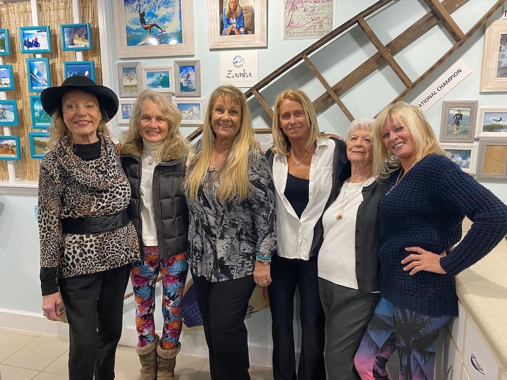 Six women posing indoors in front of wall art and a large wooden ladder display.
