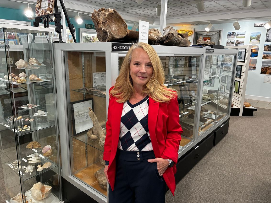 Woman in red blazer standing in a museum exhibit room beside glass display cases and animal specimens