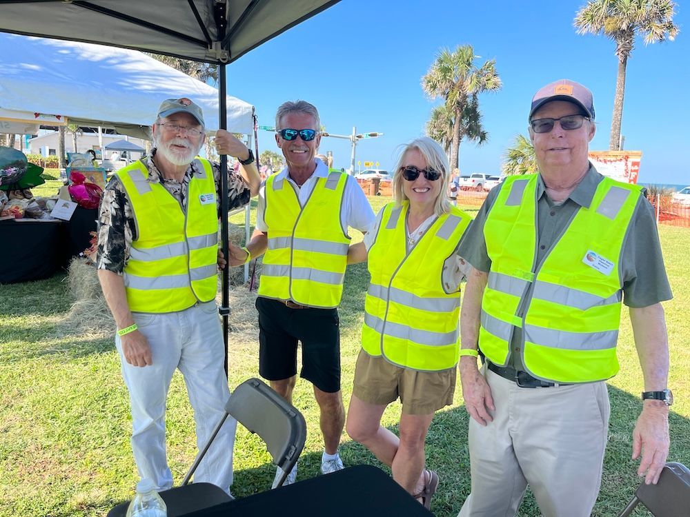 Four people in neon safety vests stand outdoors near a tent on a sunny day.