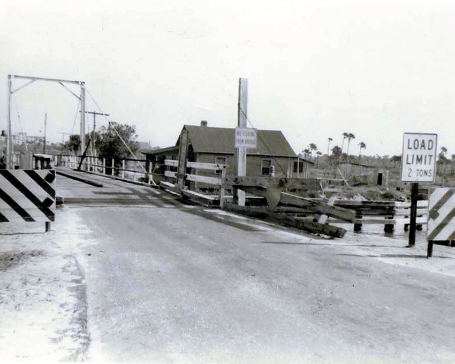 Black-and-white view of a drawbridge with a load limit sign and a small house beyond the bridge