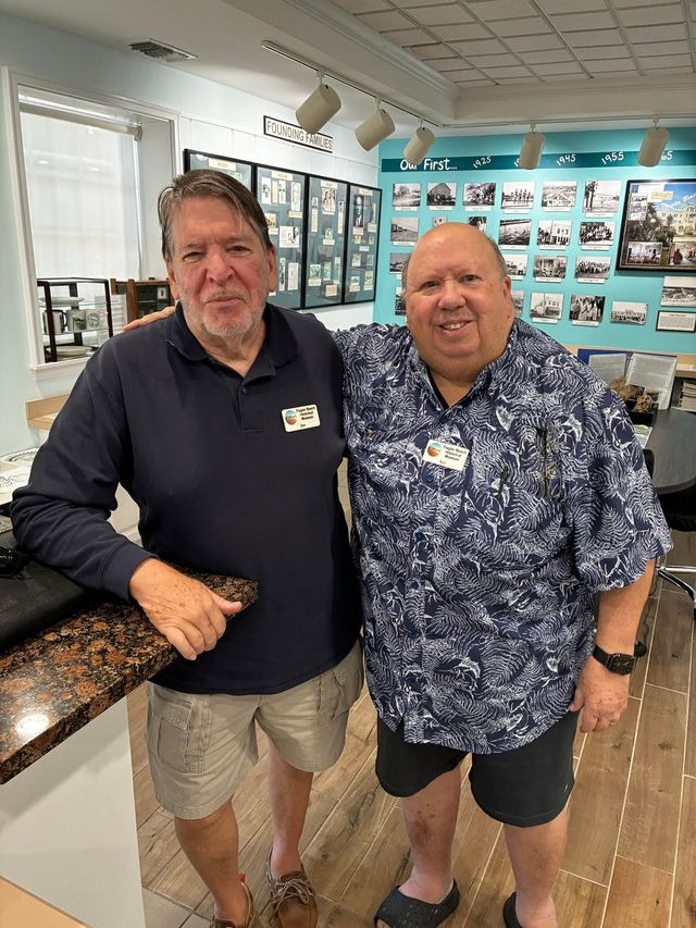 Two men smiling indoors at a counter, with framed photos displayed on the wall behind them.