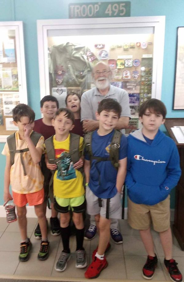 Five boys with backpacks standing in front of a scout troop display case and “Troop 495” sign