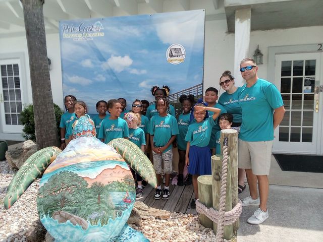 Group posing with a large sea turtle sculpture in front of a building and ocean-themed backdrop.