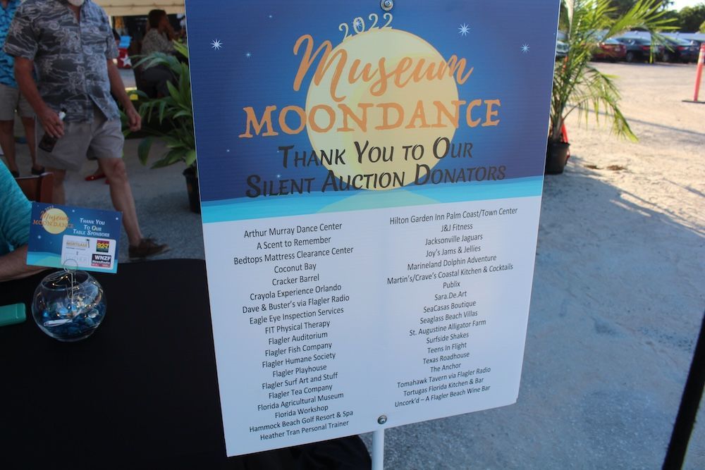 Museum Moondance silent auction sign on a blue stand outdoors, with people and items nearby