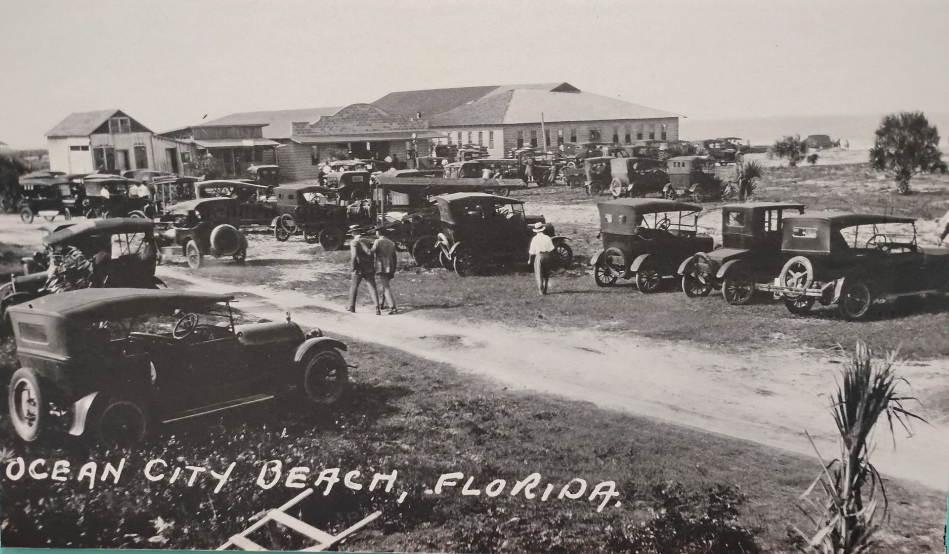 Vintage black-and-white photo of Ocean City Beach, Florida, with early cars, people, and beach buildings.