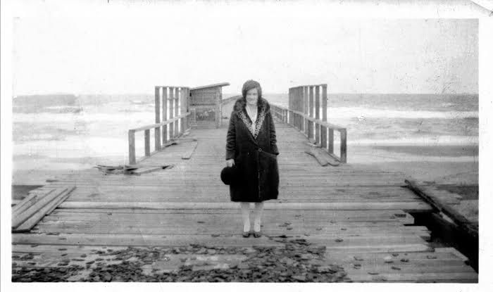 Woman standing on a wooden pier in a black coat, facing the camera with the sea behind her.