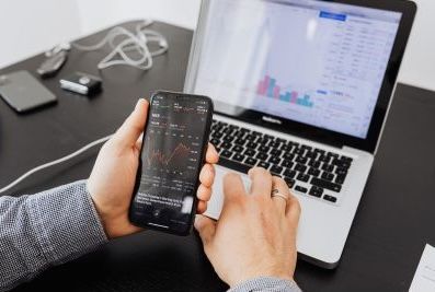 Hands using a smartphone beside a laptop displaying charts and code on a desk