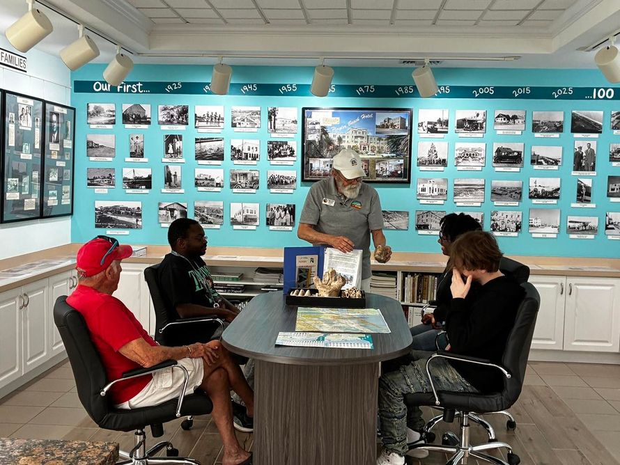 Four people sit around a table in a bright museum exhibit room with photo displays on turquoise walls.