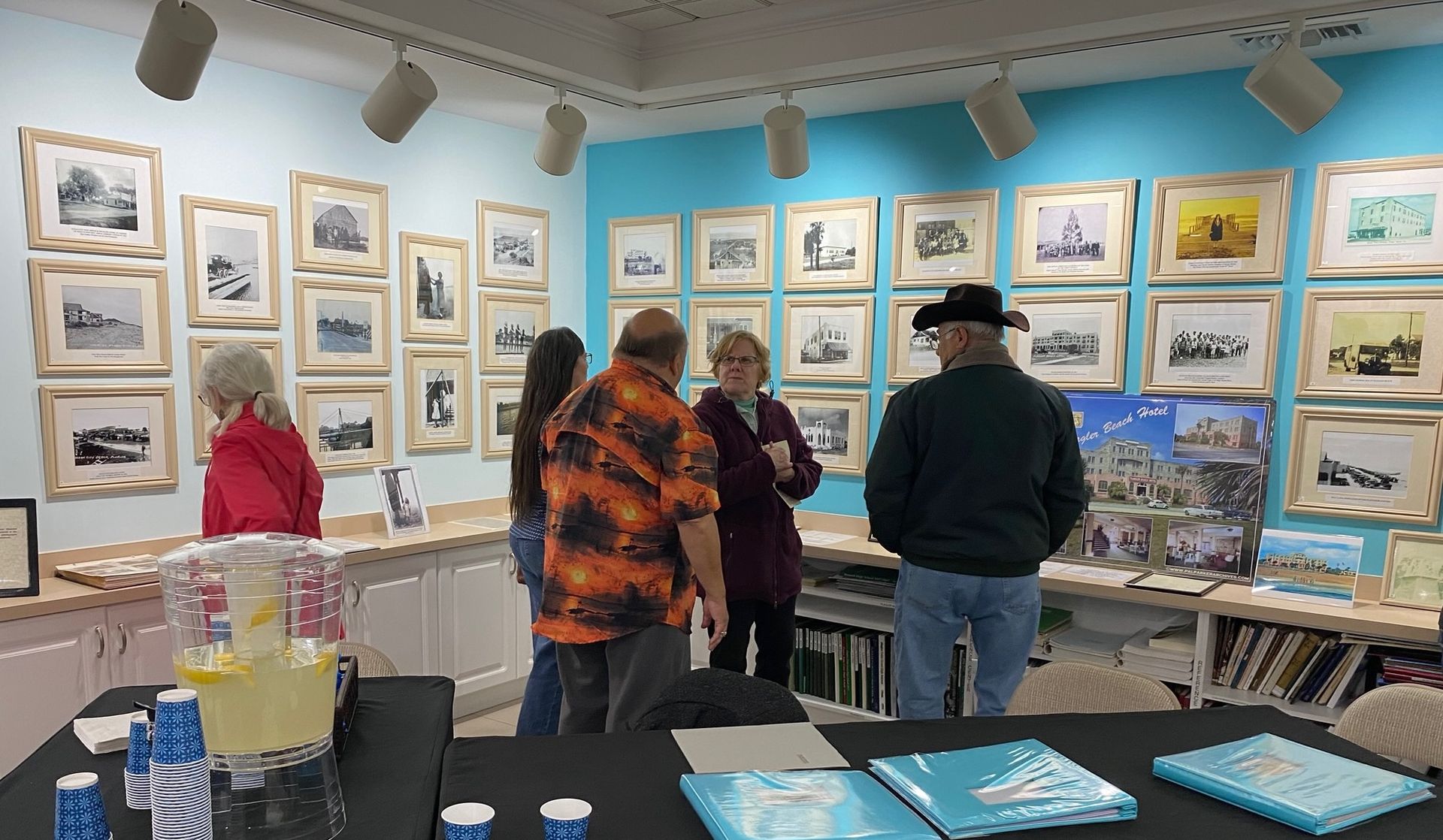 Art gallery with framed paintings on blue walls; a few visitors browse prints and books.