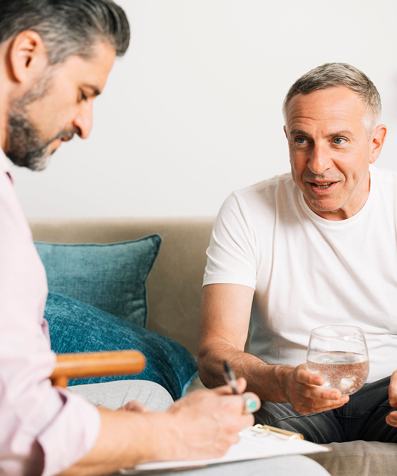 Two men are sitting on a couch talking to each other and one is holding a glass of water.