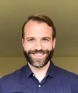 A man with a beard is smiling for the camera while wearing a blue shirt.