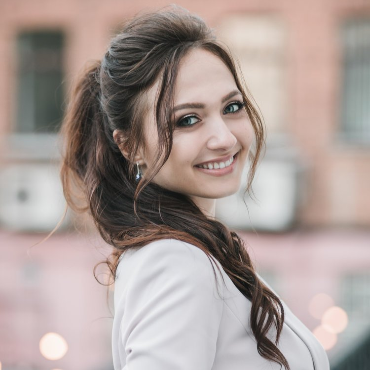 Woman with brown hair in a ponytail, smiling, wearing a blazer, outside near a building.