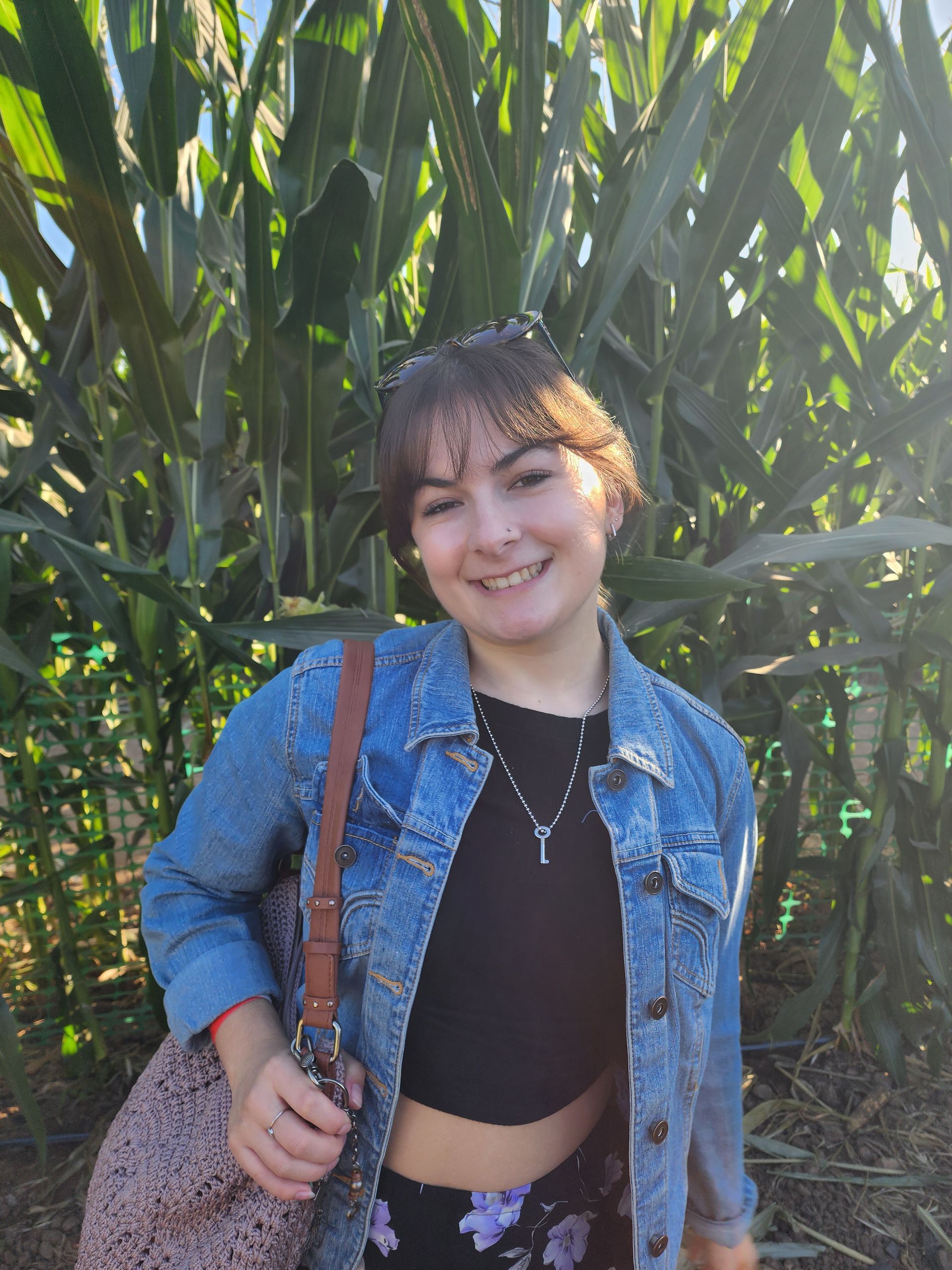 Woman with short brown hair smiles in a corn maze, wearing a jean jacket, black top, and carrying a patterned bag.