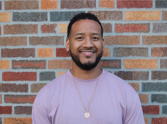 Smiling man in a pink shirt, gold necklace, stands against a brick wall.