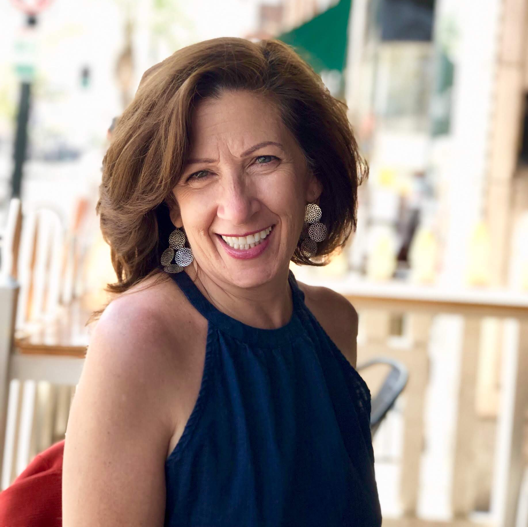 Woman with brown hair, smiling, wearing a dark blue halter top, outdoor cafe setting.