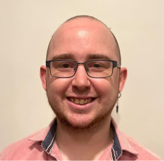 Man with glasses smiling, wearing a pink shirt, small beard, and dangling earring.
