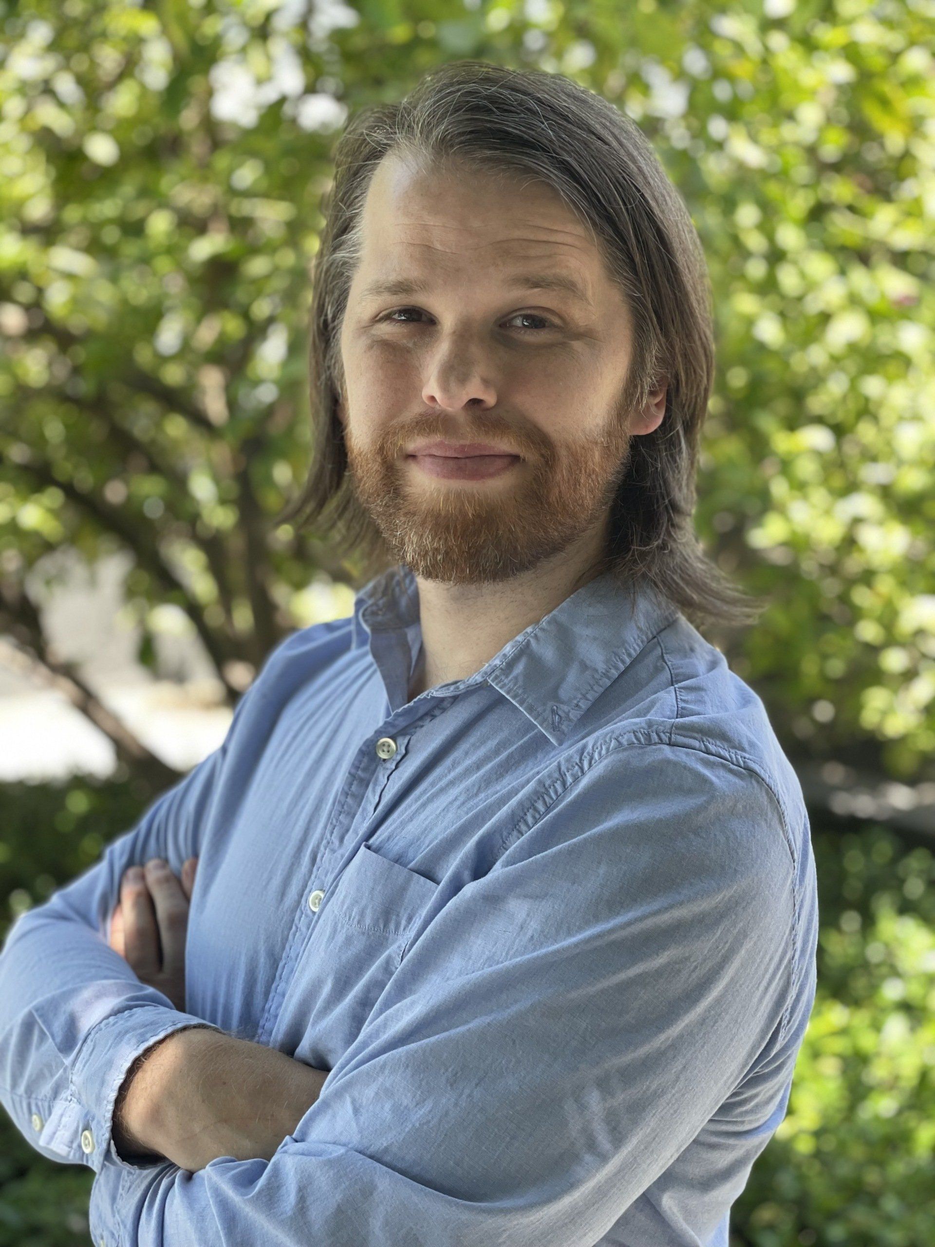 Man with brown beard and long brown hair, arms crossed, wearing a blue button-down shirt, in front of greenery.