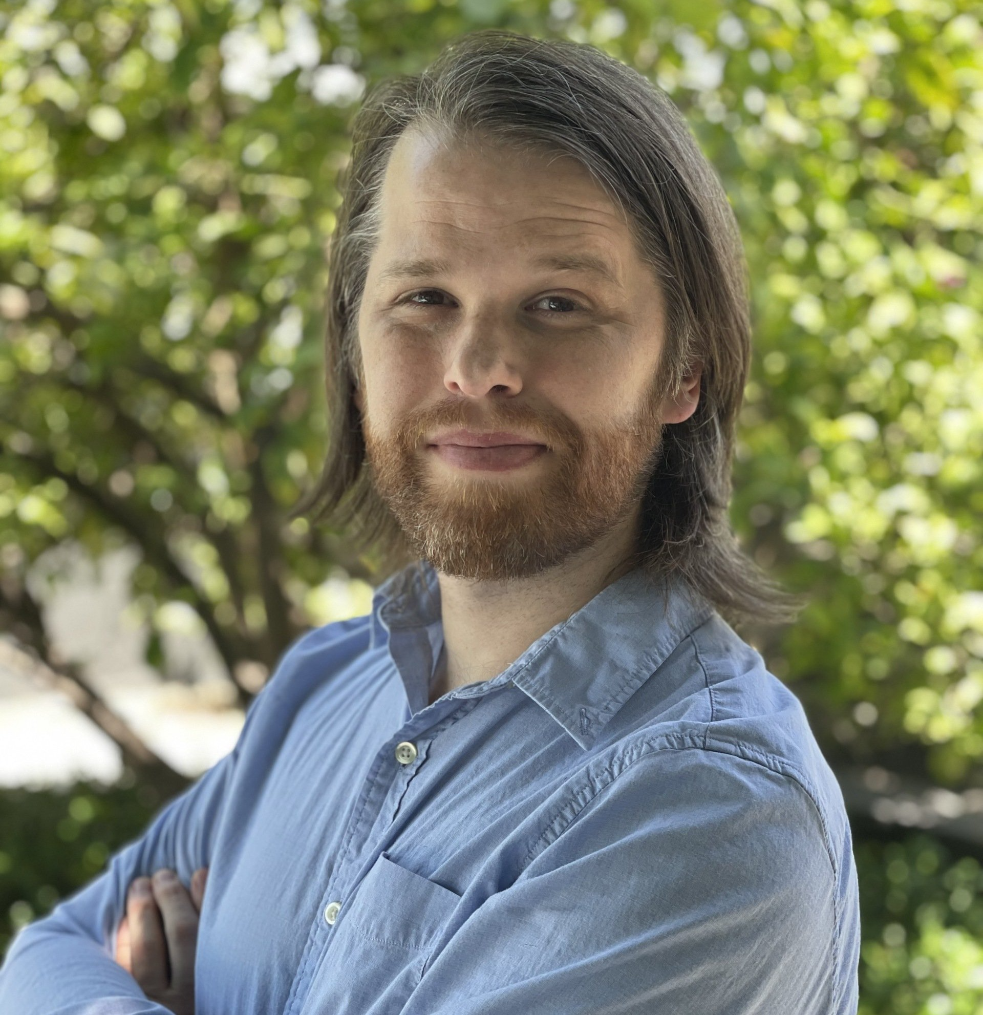 Man with beard and long hair, arms crossed, smiles at camera, blue shirt, outdoor setting.