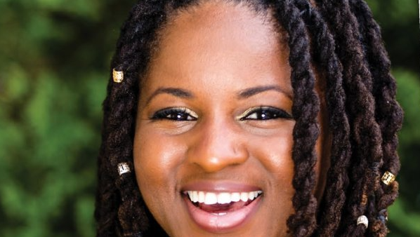 Smiling Black woman with locs, wearing gold eyeshadow, against a blurred green background.