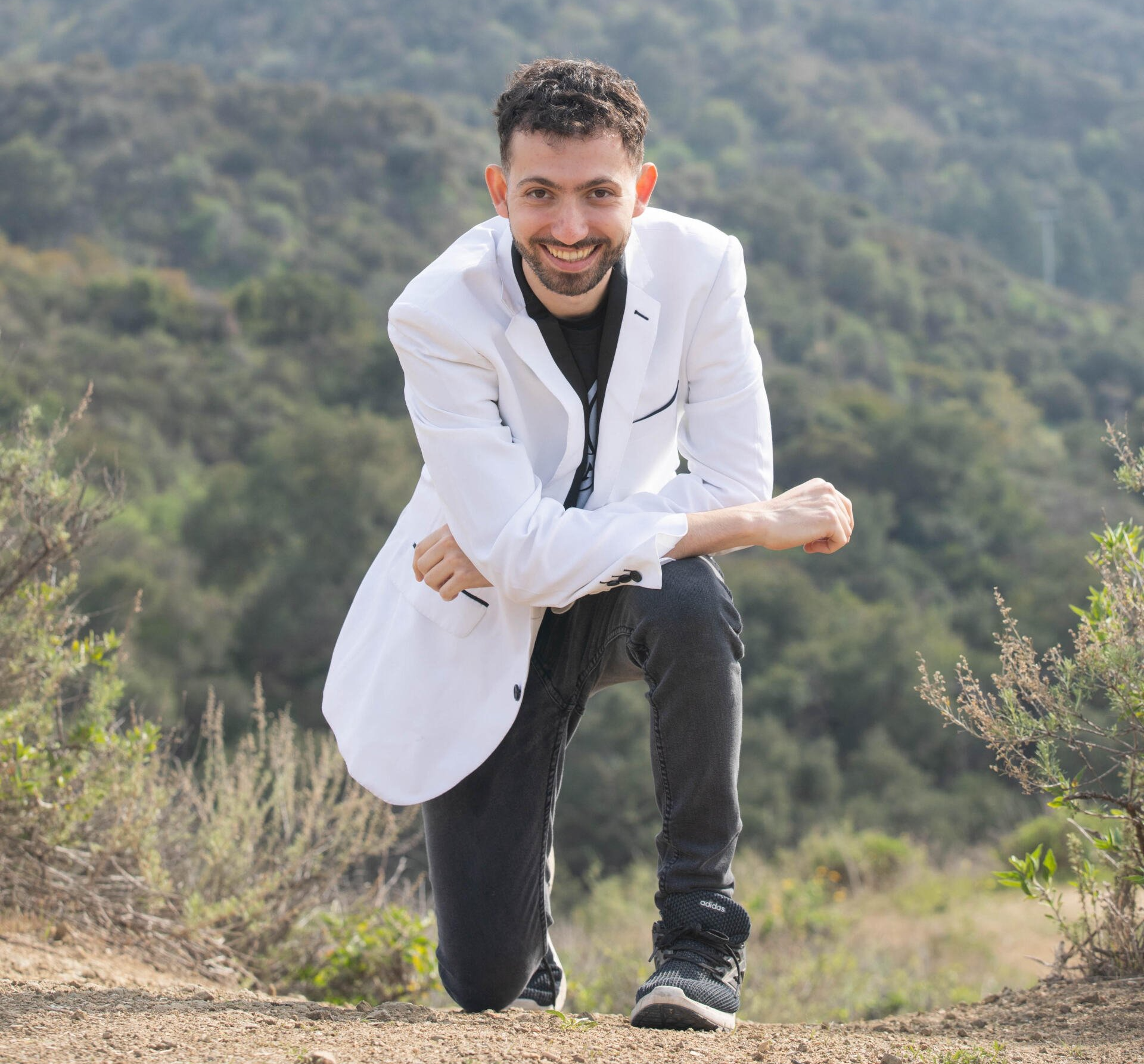Man kneels on a dirt path in nature, wearing a white coat, smiling.