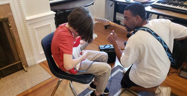 A guitar instructor kneels, guiding a student playing guitar. Indoors, a red shirt, black amplifier, and keyboards.
