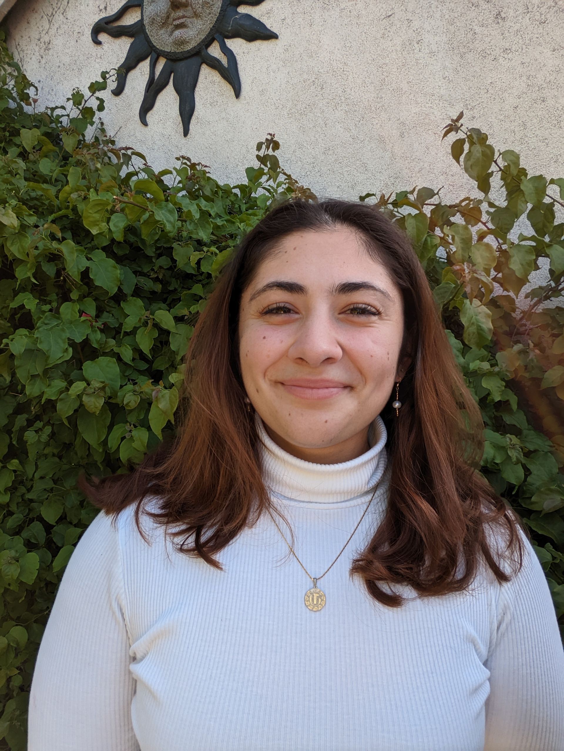 Woman with brown hair and a white turtleneck smiles, against a backdrop of vines and a sun-shaped wall decoration.