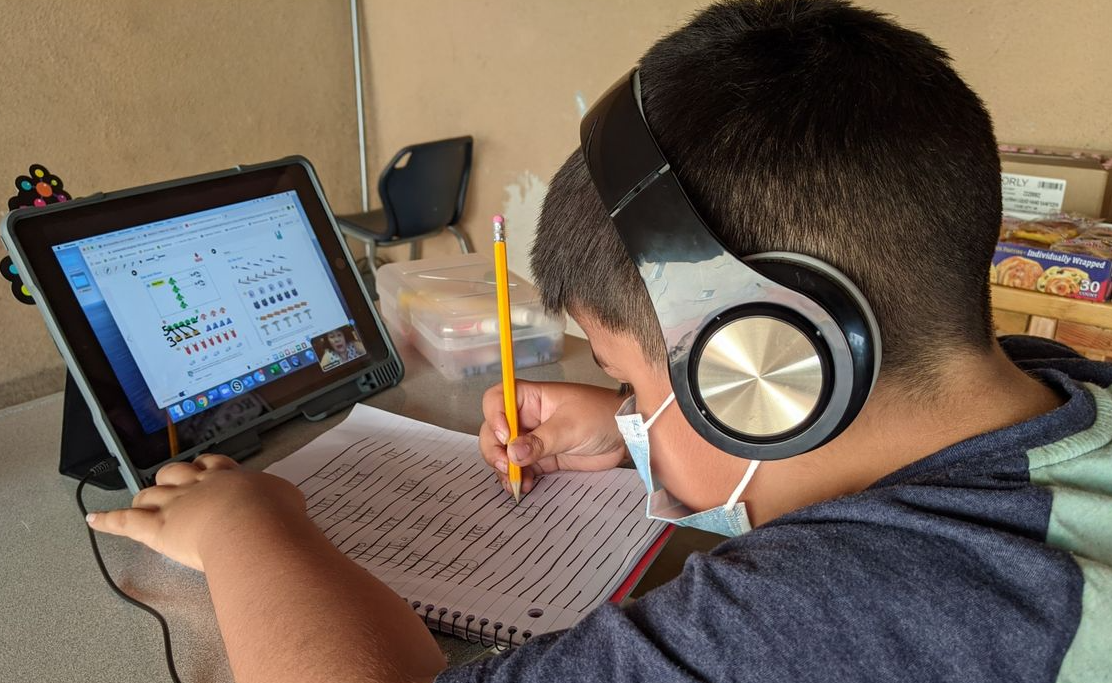 A child with headphones and a mask studies at a tablet, writing in a notebook.