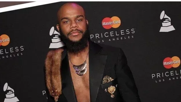 Man with a beard and fur stole poses at a red carpet event.