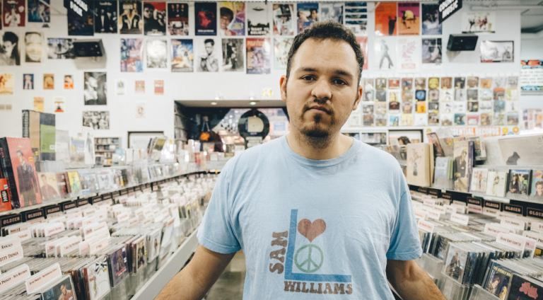 Man in a blue t-shirt, standing in a record store. Albums and posters are visible.