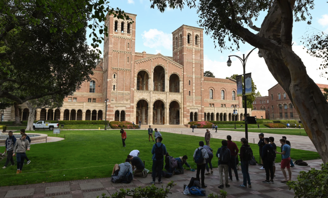 Students on a green lawn in front of a brick building with two tall towers and arched openings.
