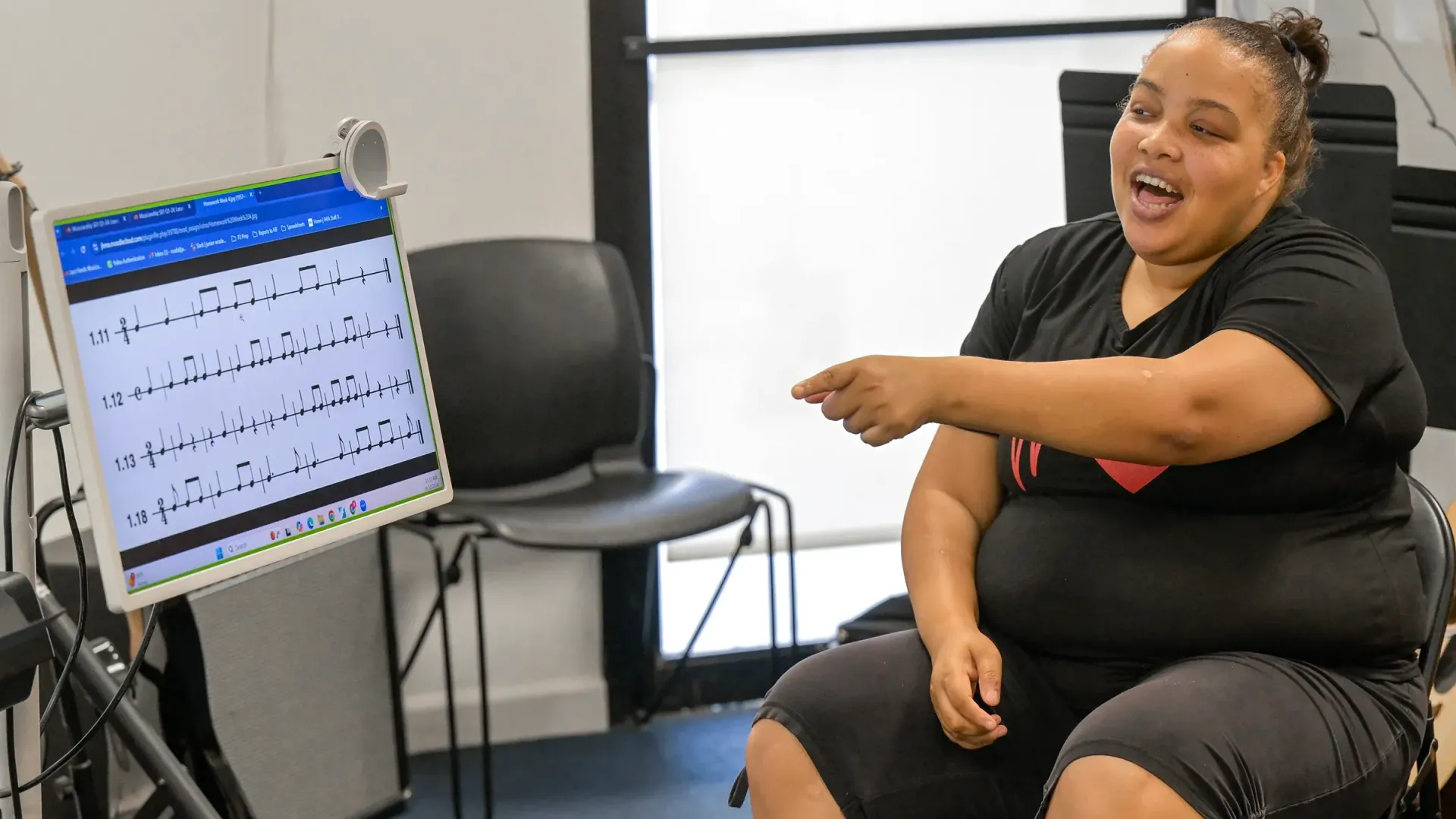 Woman singing, pointing, at a music screen in a room. She wears a black shirt and sits on a chair.