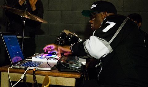 DJ in a black jersey, cap, mixing music on laptop & controllers, dark setting, hands visible.