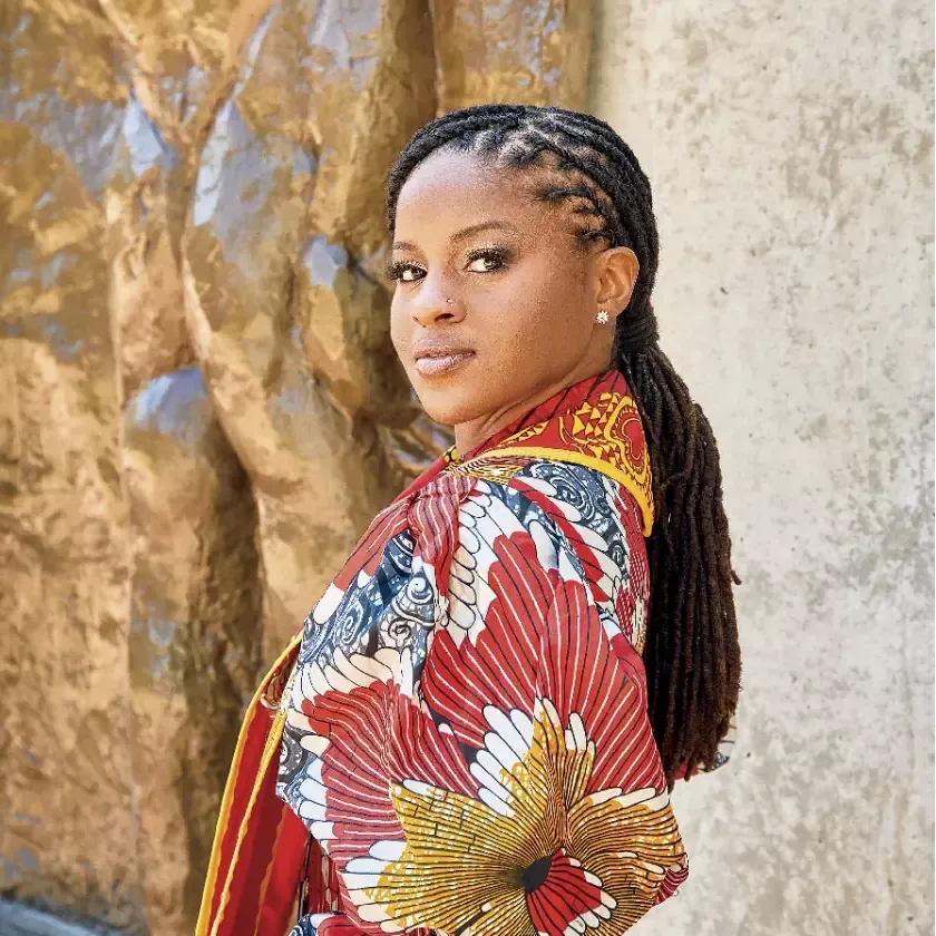 Woman with braided hair wearing a colorful patterned wrap, standing outdoors next to a textured wall.