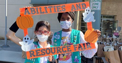 Two girls in Girl Scout vests hold a sign reading
