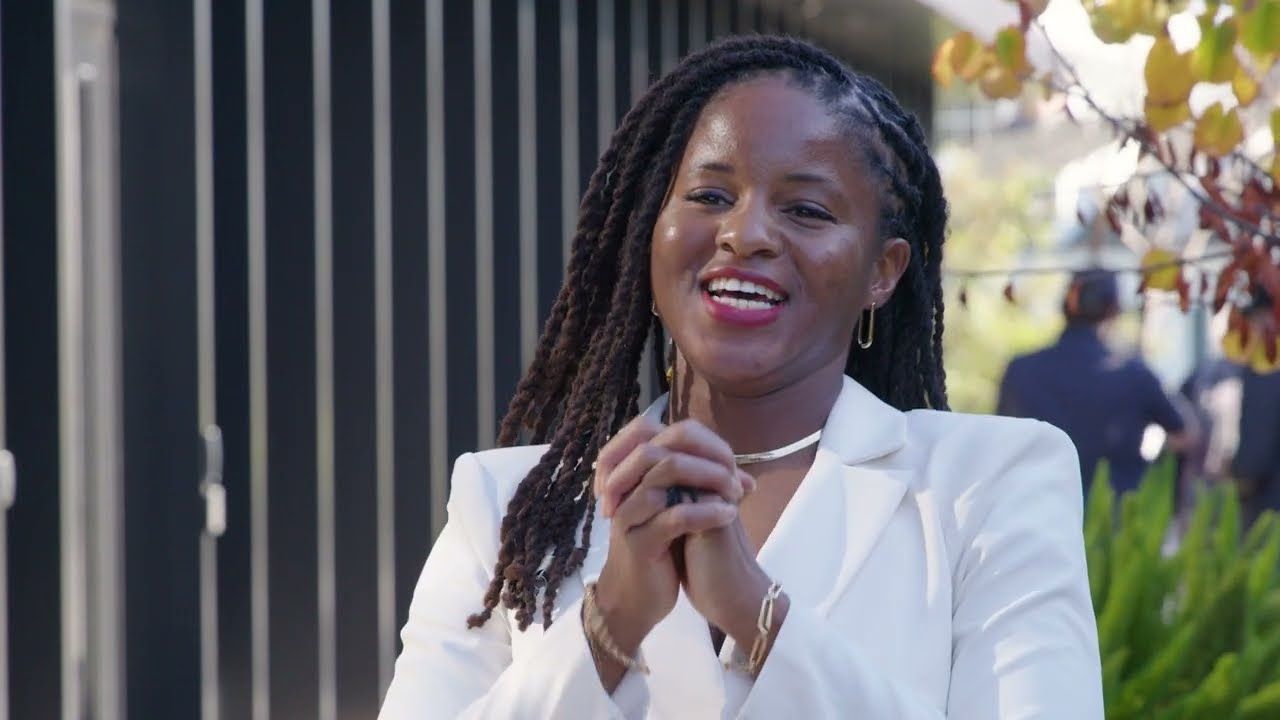 Woman with long, dark locs in a white blazer smiles outdoors, hands clasped.