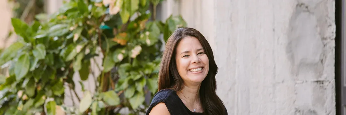 Woman with long brown hair smiling, in front of greenery and a white wall.