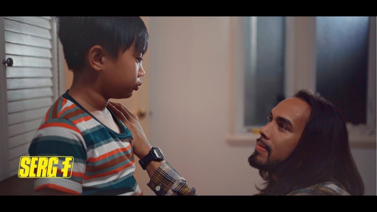A man comforts a young boy inside a room. The man has long hair and a beard, the boy is wearing a striped shirt.