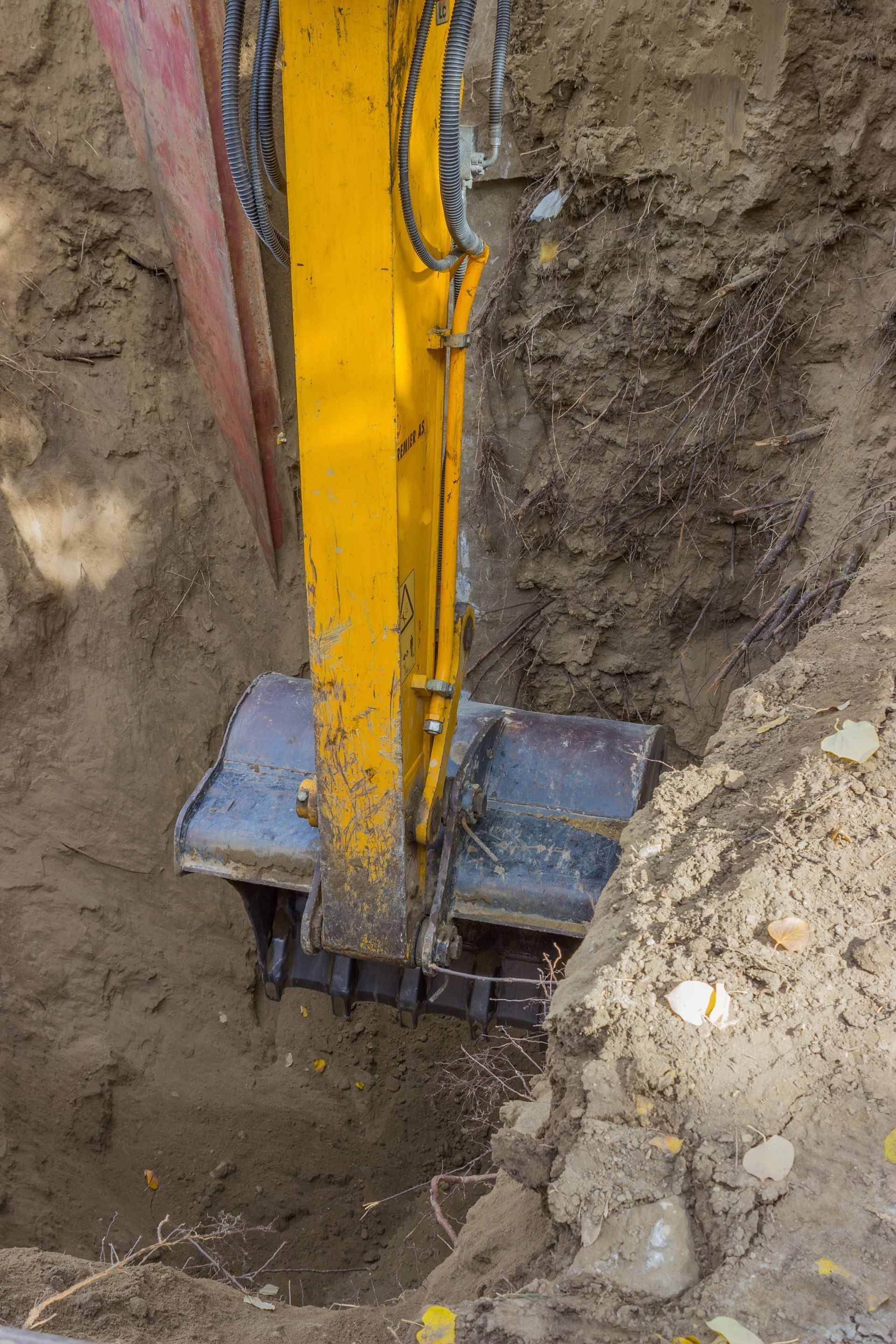Yellow excavator bucket digging in a narrow trench beside packed earth and concrete rubble