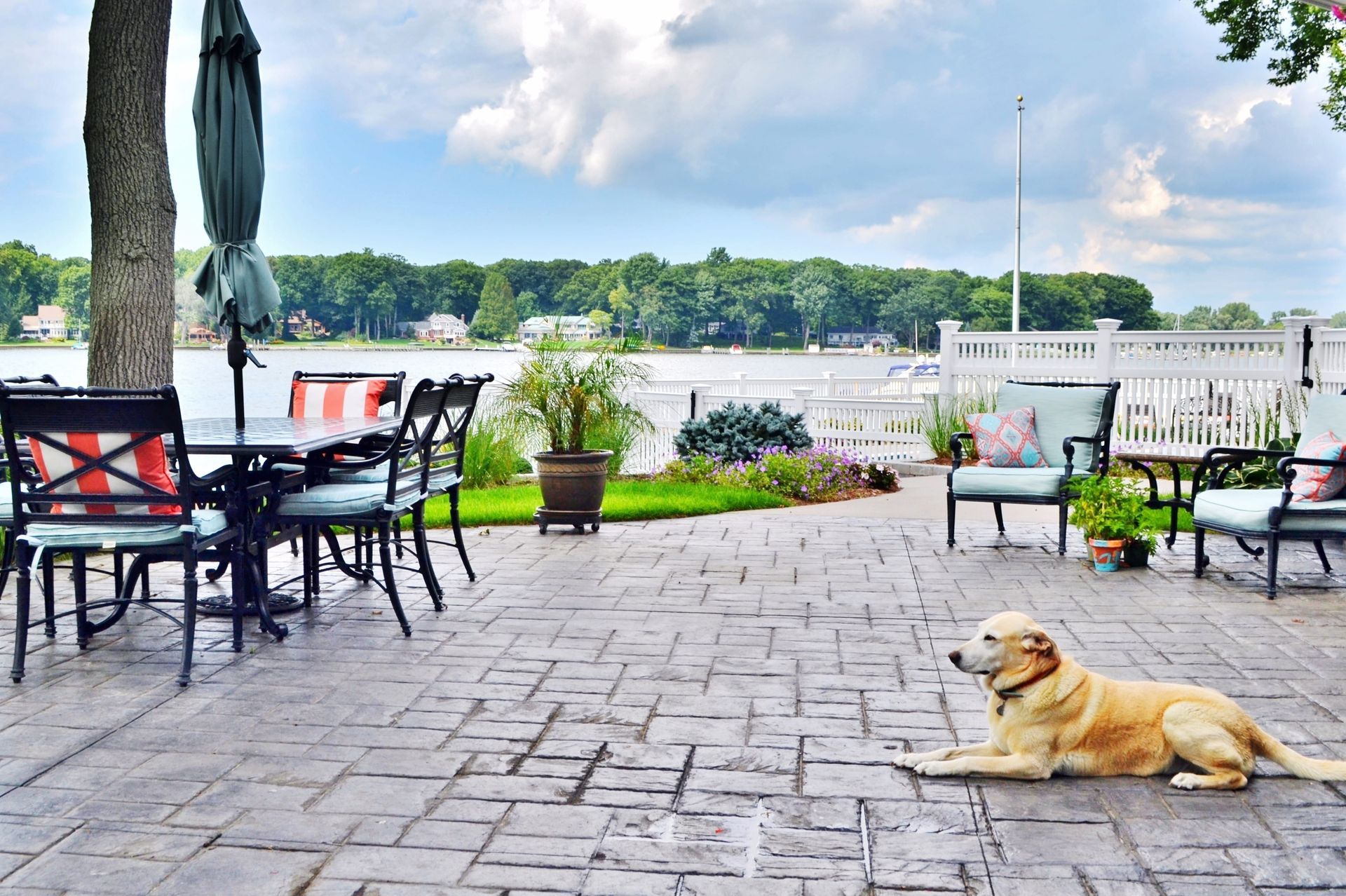 Patio overlooking a lake, with tables, chairs, and a golden dog lying on the stone ground.