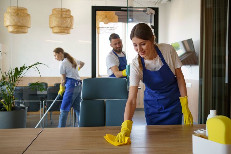 Three people wearing blue aprons clean an office. Woman wipes table, others mop and look on.