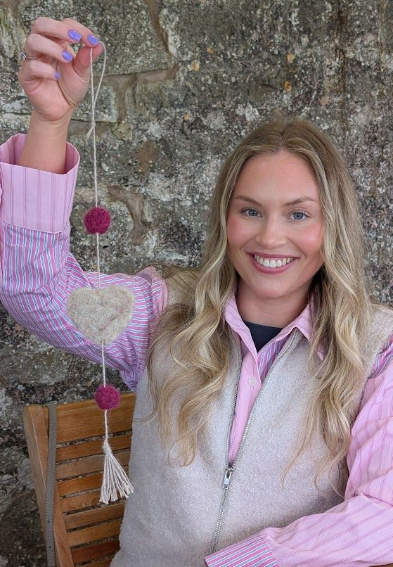 Smiling woman holds a pink heart ornament beside a tree trunk outdoors