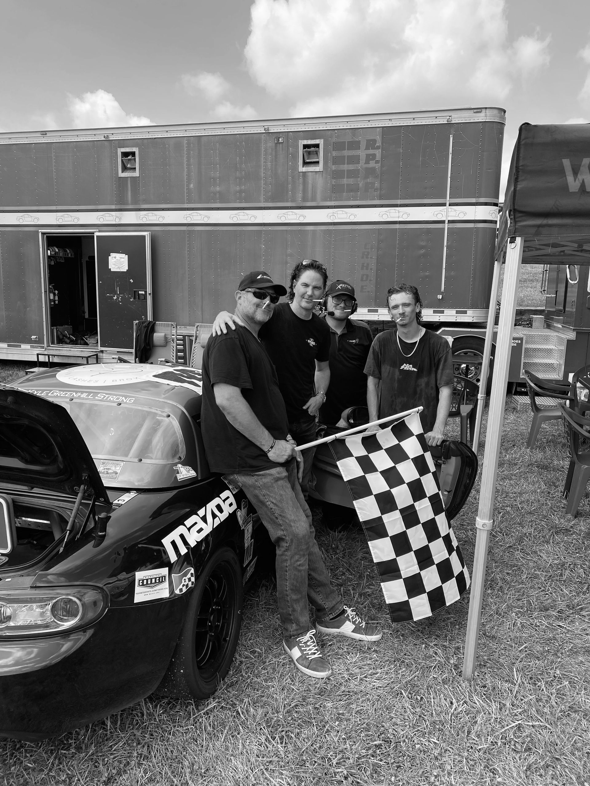 Three people posing by a black car, holding a checkered flag at an outdoor event.