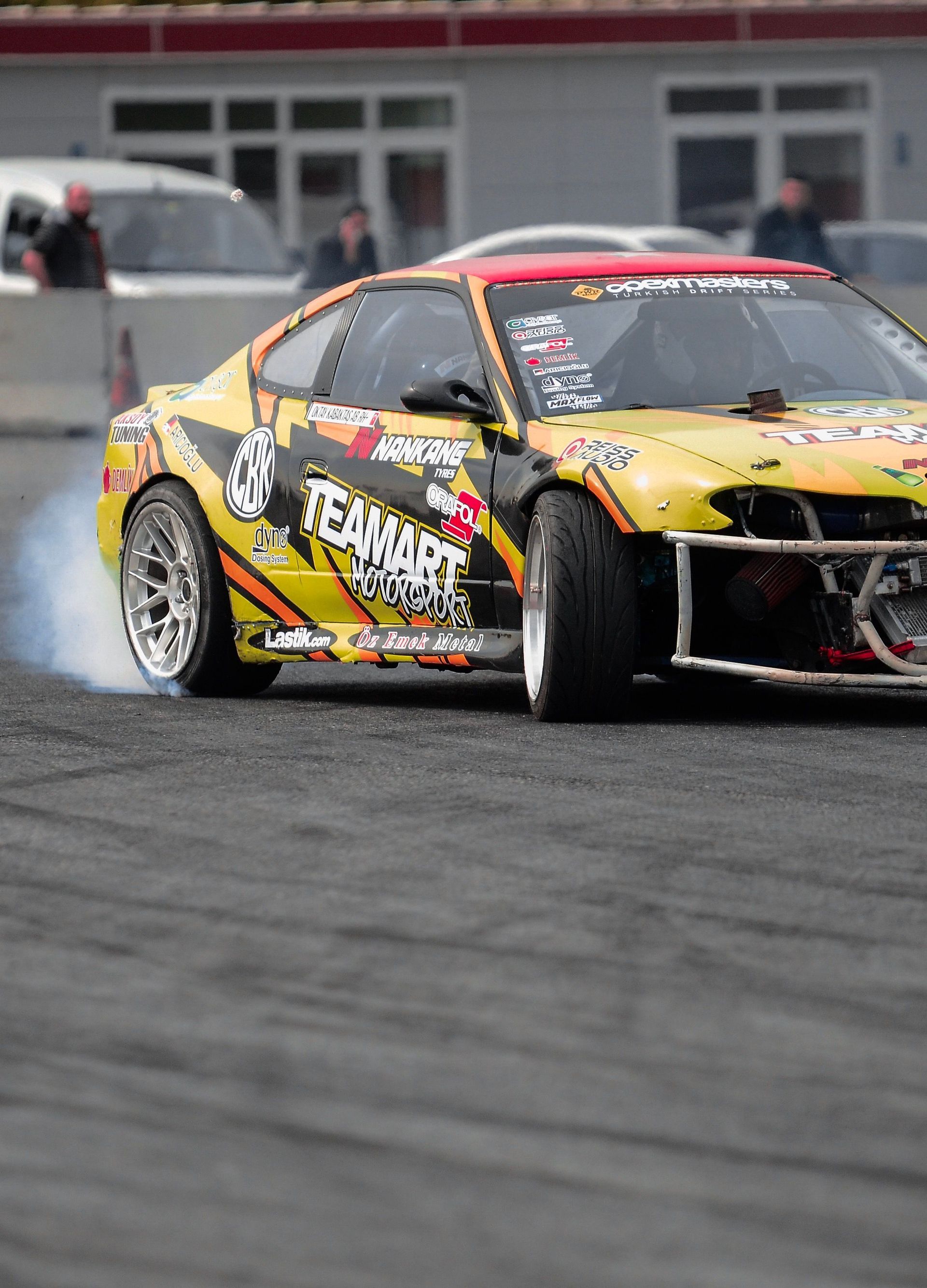 Yellow-and-black drift car sliding on a racetrack, front wheels turned sharply with smoke trailing behind.