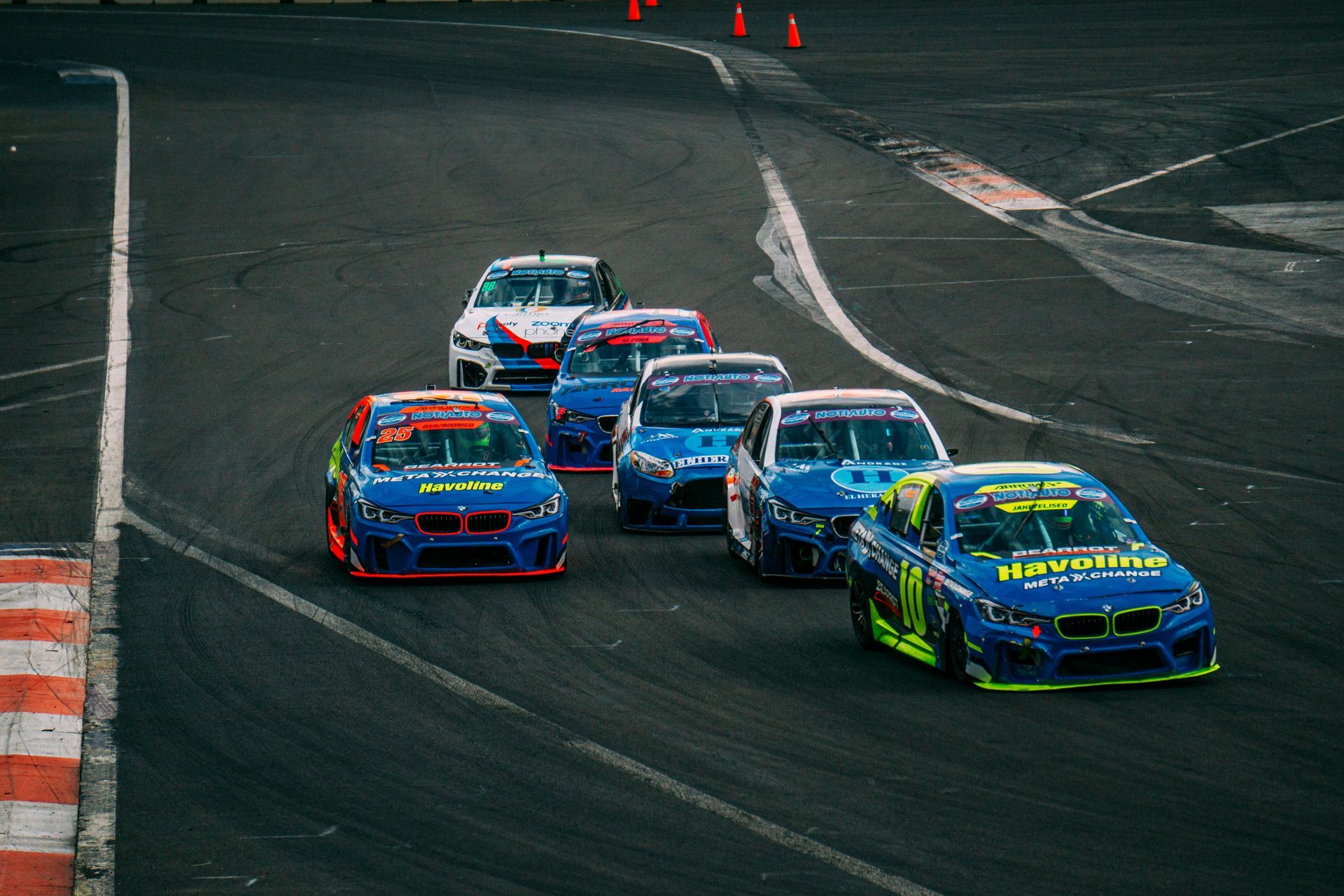 Five race cars in a tight pack turning on a dark racetrack, led by a green car