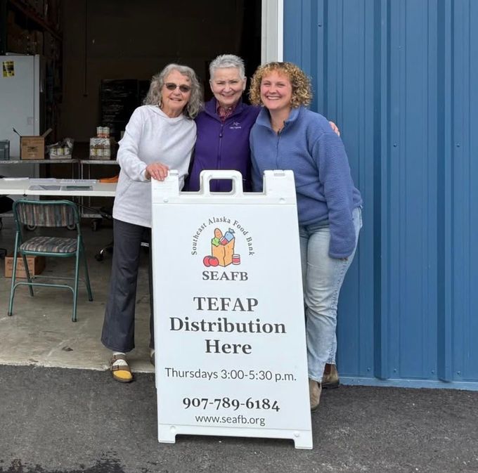 Three women stand beside a “TEFAP Distribution Here” sign outside a blue building.
