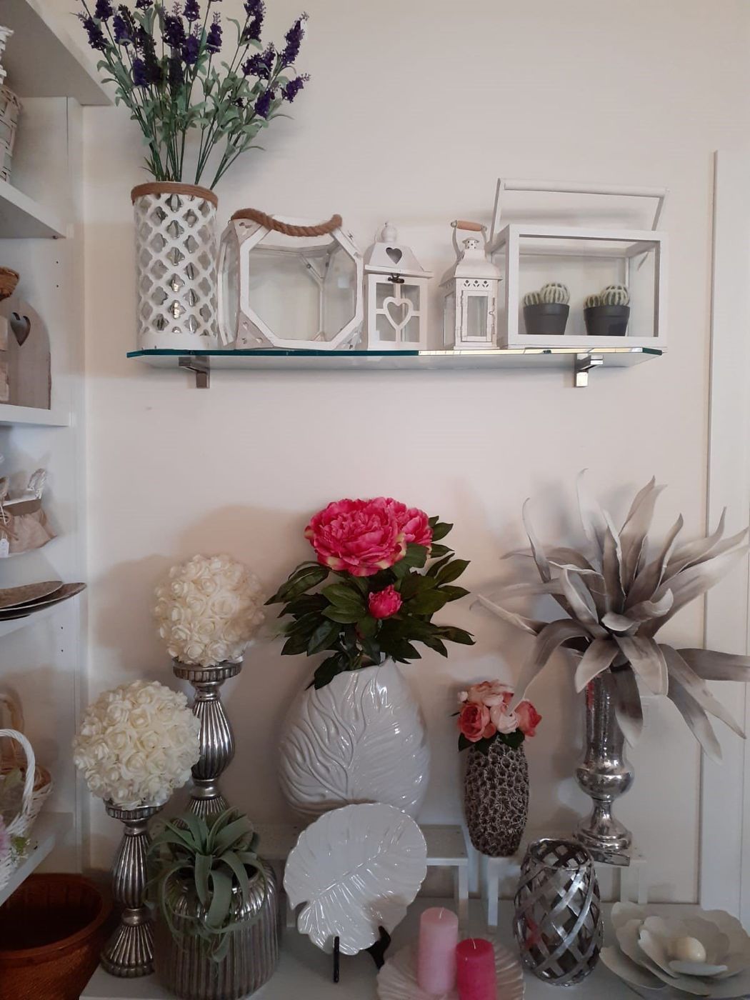 Esposizione di oggetti decorativi: fiori in vasi, candele e lanterne su scaffali bianchi.