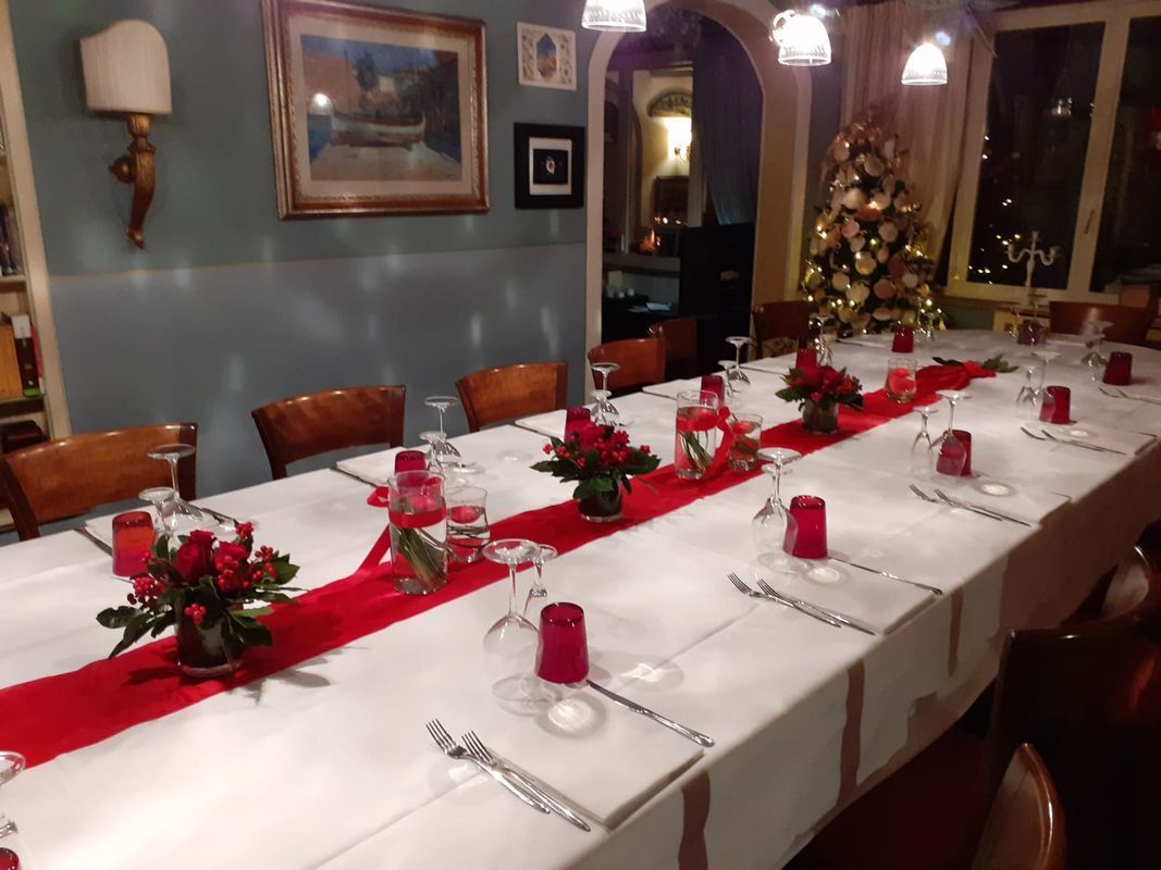 Lungo tavolo da pranzo apparecchiato per un pasto festivo, con dettagli rossi e un albero di Natale sullo sfondo.